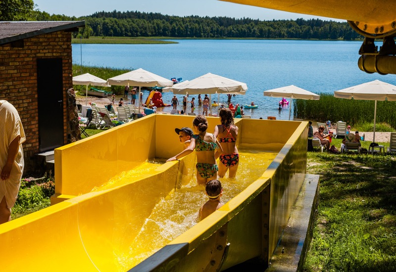 Dzieci wchodzące na jasną żółtą zjeżdżalnię wodną w ośrodku nad jeziorem; piaszczysta plaża z parasolami i leżakami