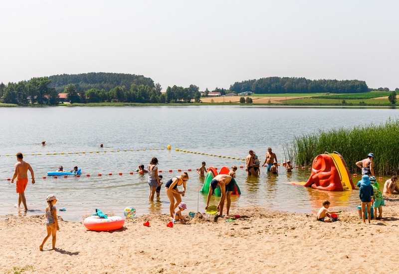 Rodzinna plaża nad jeziorem, piaszczysty brzeg, boje kąpieliskowe, bawiące się dzieci i czerwona zjeżdżalnia.