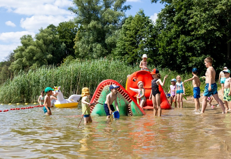 Dzieci bawią się na rodzinnym jeziornym kąpielisku w płytkiej wodzie przy kolorowej zjeżdżalni i nadmuchiwanym jednorożcu.