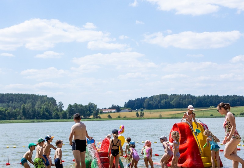 Rodziny i dzieci bawiące się na piaszczystej plaży przy jeziorze z kolorową wodną zjeżdżalnią, pod opieką dorosłych.