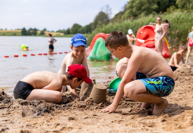 Dzieci budują zamki z piasku na rodzinnym brzegu jeziora, wydzielona strefa kąpielowa i kolorowe pedaliny w tle