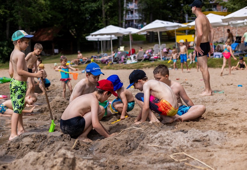 Dzieci budujące zamek z piasku na plaży hotelowej, kolorowe stroje kąpielowe, białe parasole i leżaki, przyjazne rodzinom.
