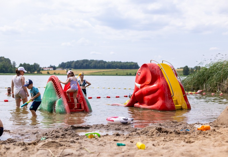 Rodzinna plaża nad jeziorem z płytką wodą i kolorową zjeżdżalnią w kształcie smoka i słonia, dzieci bawią się, rodzice brodzą
