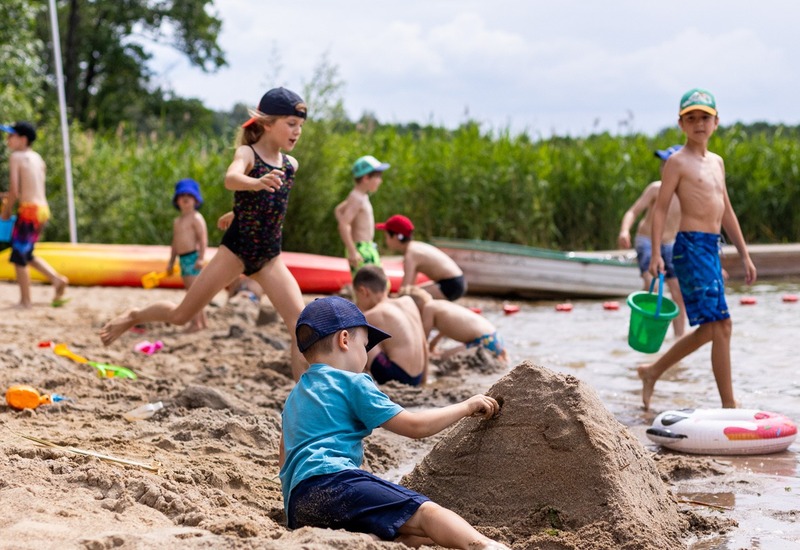 Dzieci na rodzinnej plaży nad jeziorem, budują zamki z piasku obok kolorowych kajaków i płytkiej wody