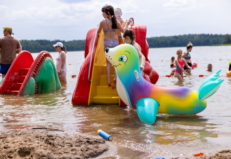 Rodzinna plaża nad jeziorem z kolorową dmuchaną foką, zjeżdżalnią wodną i dziećmi bawiącymi się w płytkiej wodzie.
