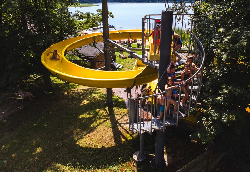 Nad jeziorem żółta spiralna zjeżdżalnia wodna, dzieci na schodach, leżaki i widok na spokojne jezioro.