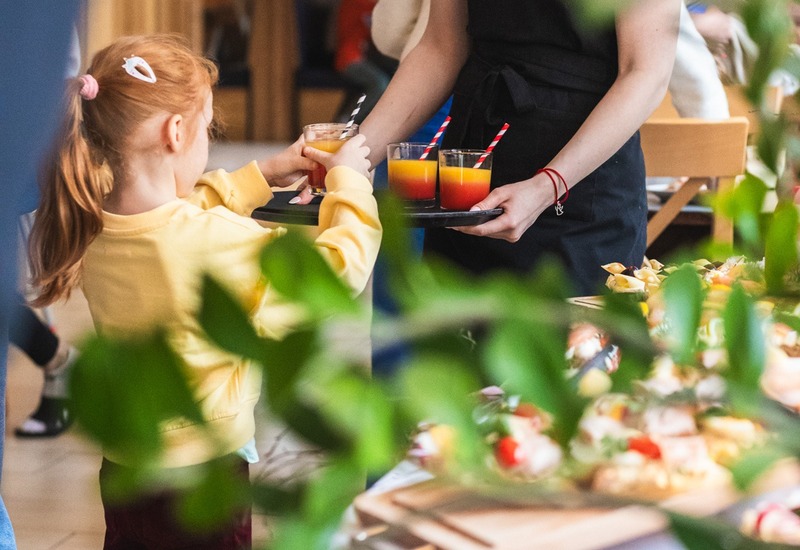 Młoda kelnerka podaje dziecku kolorowe soki na tacy przy rodzinnym bufecie w hotelowej restauracji