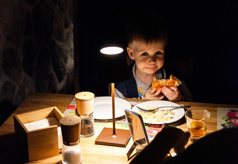 Dziecko jedzące pizzę w przytulnej, słabo oświetlonej kabinie restauracji z lampką na stoliku, talerzami i przyprawnikami