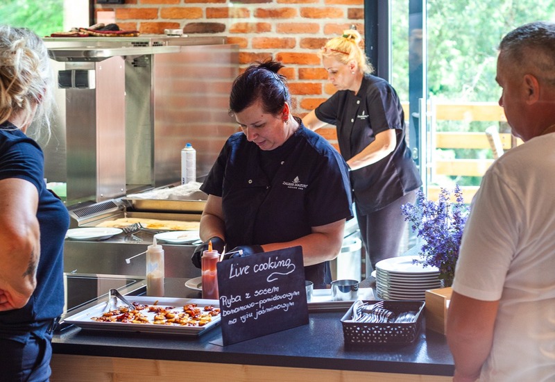 Szefowie kuchni przy otwartej stacji „Live cooking” przygotowują potrawy przy ladzie, ceglana ściana i widok na zieleń.