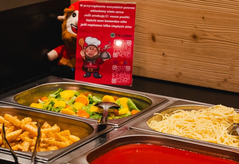 Bufet na ciepło z półmiskami: zupa pomidorowa, spaghetti, mieszane warzywa i falowane frytki na tle drewnianej ściany.