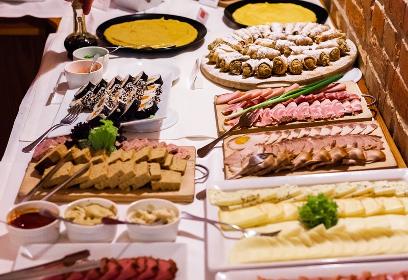 Bufet hotelowy z wędlinami, serami i ciastkami przy ceglanej ścianie; gość nalewa sos.
