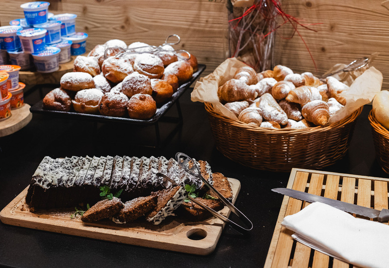 Bufet śniadaniowy: pączki z cukrem pudrem, mini rogaliki w wiklinowym koszu, krojone ciasto czekoladowe i jogurty.