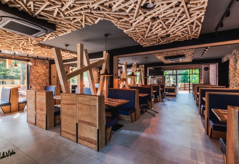 Restauracja hotelowa w stylu rustykalno‑nowoczesnym z drewnianymi boksami, dekoracyjnym sufitem z listew, lampami i oknami z widokiem na las
