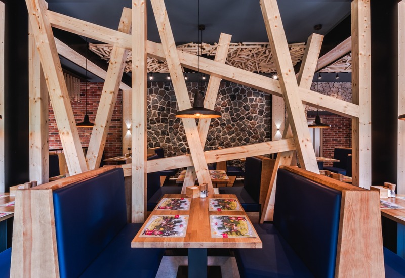 Rustykalna restauracja hotelowa z krzyżującymi się sosnowymi belkami, kamienną i ceglanymi ścianami, przytulnymi boksami i ciepłym oświetleniem.