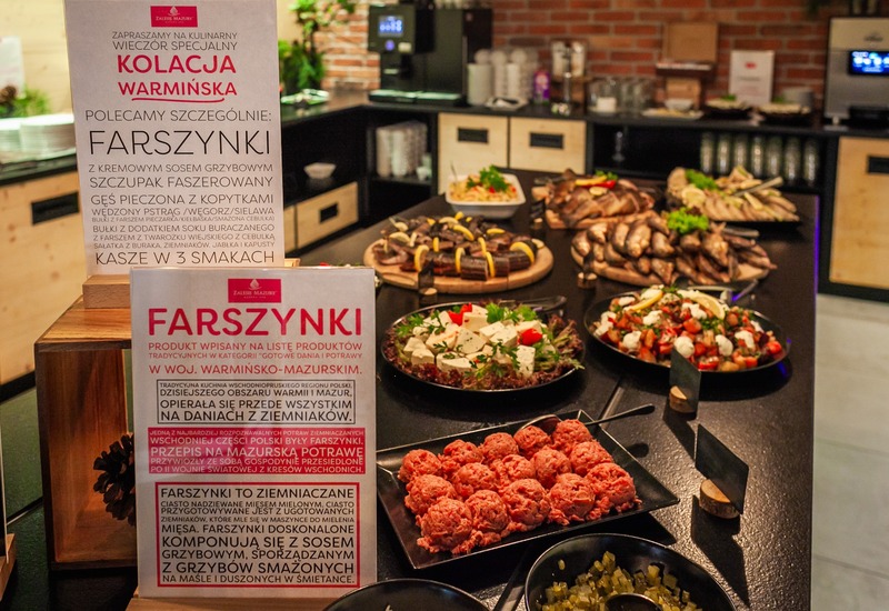Rustykalny bufet hotelowy z napisem 'Kolacja Warmińska', półmiskami wędzonych ryb, sałatek, farszynków ziemniaczanych i dodatków