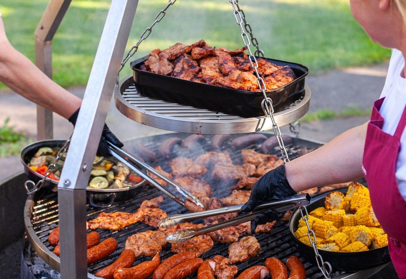 Wielopoziomowy grill na zewnątrz, personel grilluje kiełbasy, kurczaka i kukurydzę na zawieszonym ruszcie podczas hotelowego grilla