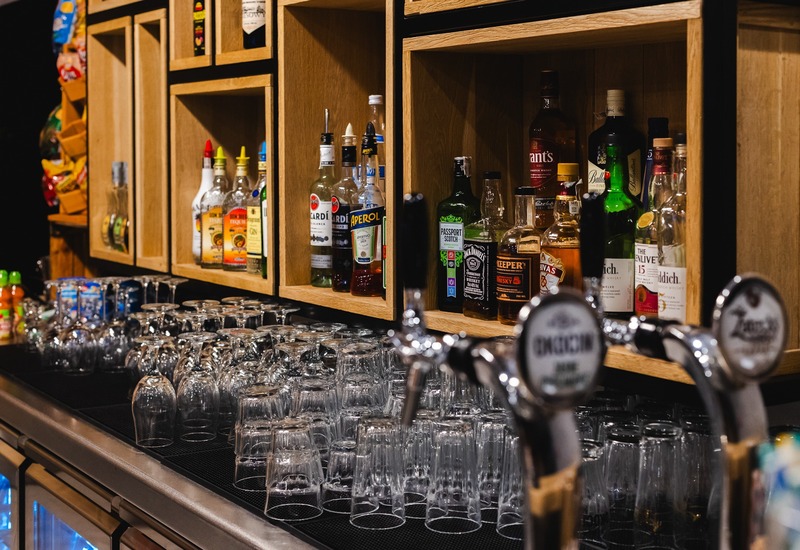 Bar hotelowy z drewnianymi półkami pełnymi alkoholi, krany piwne na pierwszym planie i rzędem czystych kieliszków.
