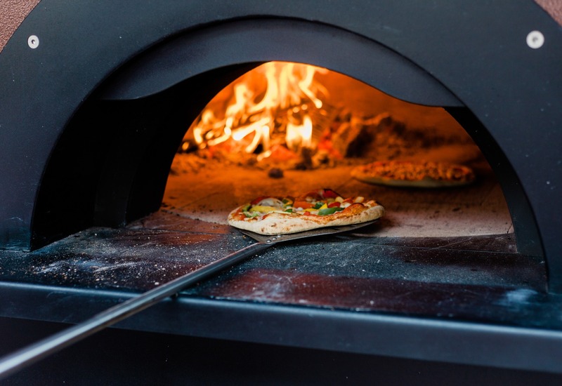 Piec opalany drewnem z żywym ogniem i świeżo upieczoną pizzą na łopacie, podkreślający ofertę restauracji hotelowej.
