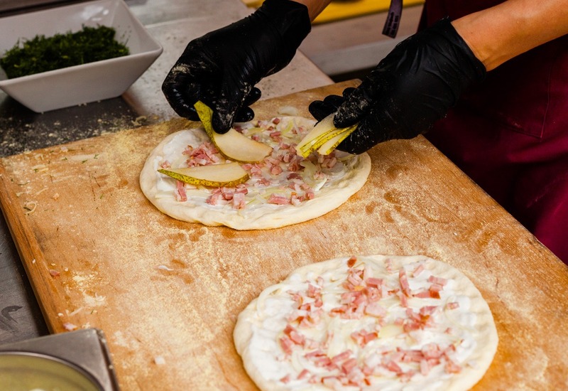 Kucharz przygotowuje rzemieślniczą pizzę w hotelowej kuchni, w czarnych rękawiczkach układa plastry gruszki na kremie z szynką i cebulą.