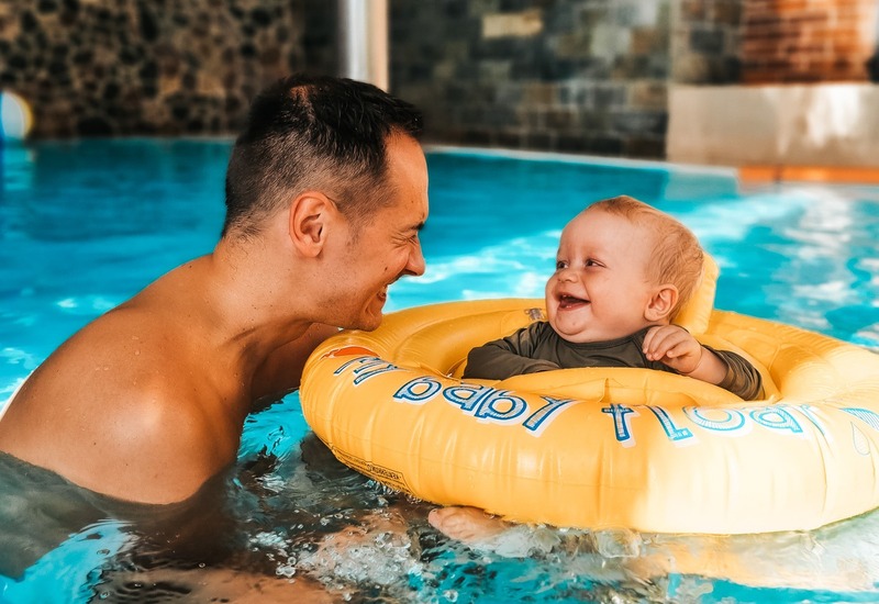 Ojciec bawi się z uśmiechniętym niemowlęciem w żółtym kółku na krytym basenie hotelowym z ciepłymi ceglanymi ścianami.