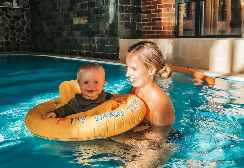 Mama i niemowlę w krytym basenie hotelowym, dziecko w żółtym kółku do pływania, ceglana ściana, duże okno i słoneczna tafla wody