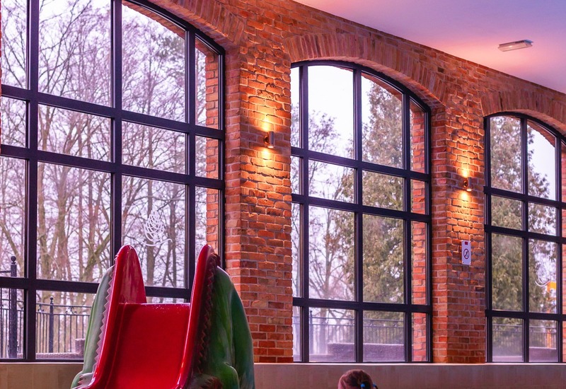 Kryty basen hotelowy z wysokimi, łukowymi oknami z cegły, czerwoną zjeżdżalnią i dziećmi bawiącymi się w wodzie.