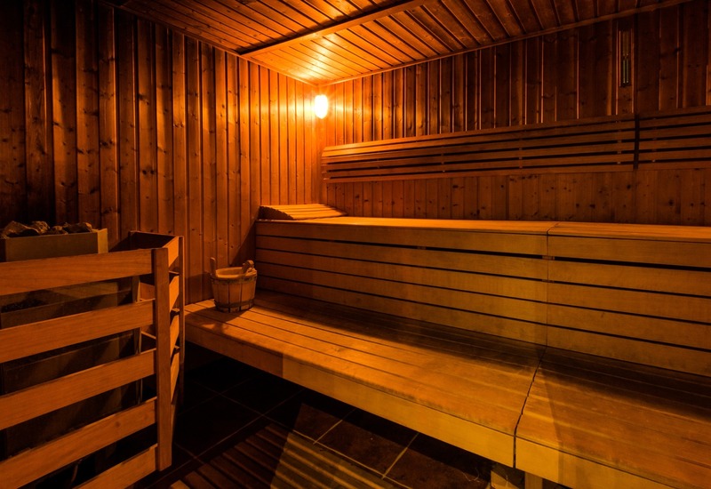 Sauna hotelowa obita drewnem, piętrowe ławy, piec z kamieniami i wiadro, łagodne bursztynowe światło, relaks w spa.