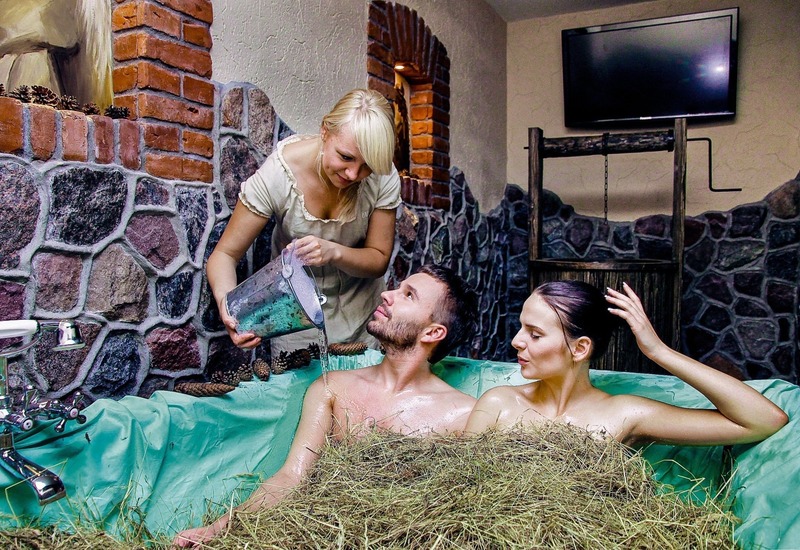 Para korzysta z tradycyjnej kąpieli w sianie w rustykalnym spa; obsługa wlewa wodę przy kamienno‑ceglanych ścianach
