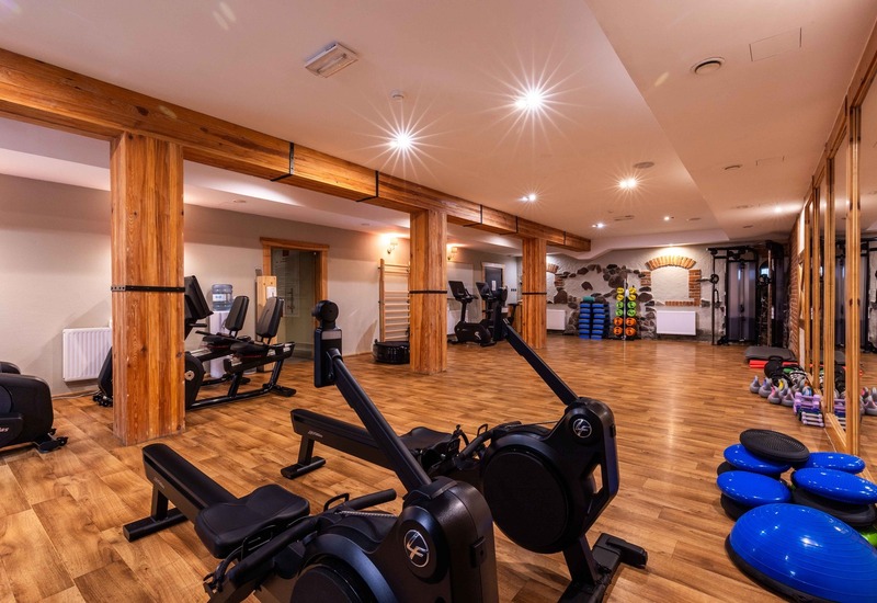Przestronna sala fitness w hotelu z drewnianymi belkami, maszynami wioślarskimi, rowerami, ciężarkami i materacami.