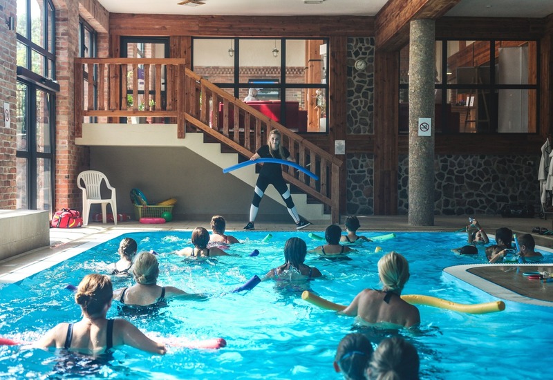 Kryty basen hotelowy z aqua aerobikiem: instruktorka na brzegu z niebieskim makaronem prowadzi ćwiczenia w przytulnej, drewniano-kamiennej przestrzeni.