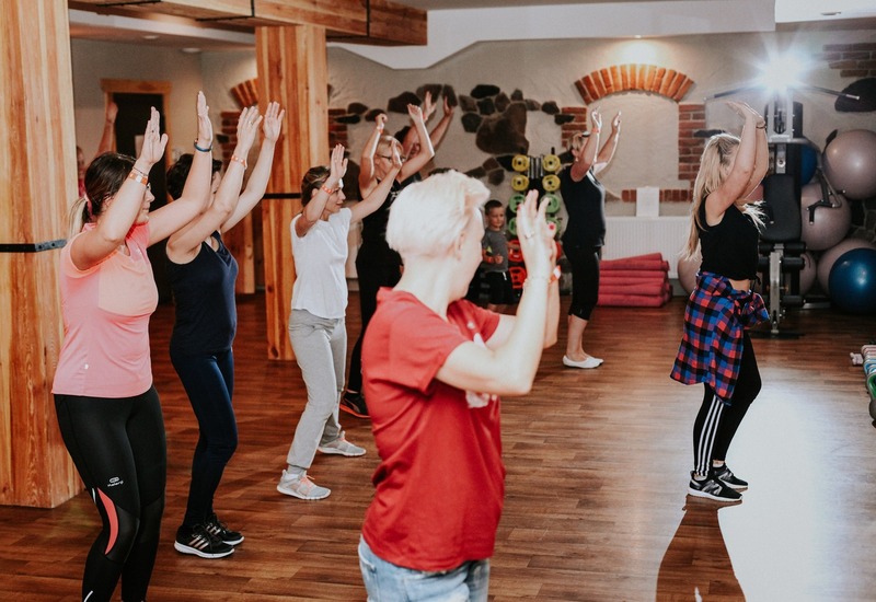 Zajęcia fitness w hotelowym studio — instruktorka prowadzi uczestników unoszących ręce w przytulnym, drewnianym pomieszczeniu z piłkami i ciężarkami
