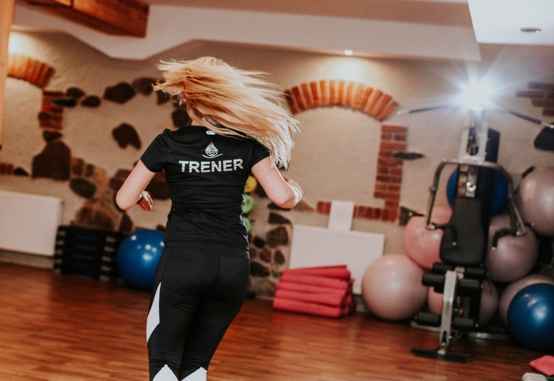 Trenerka w hotelowym studiu fitness prowadzi energetyczne zajęcia; drewniana podłoga, piłki i maty przy kamienno-ceglanych ścianach.