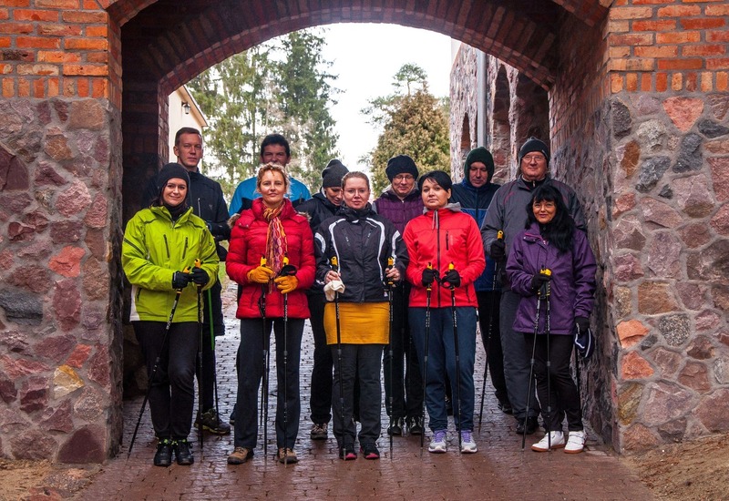 Goście hotelowi w kolorowych kurtkach trzymają kijki do nordic walking pod kamienną arkadą, gotowi na prowadzony, aktywny spacer.