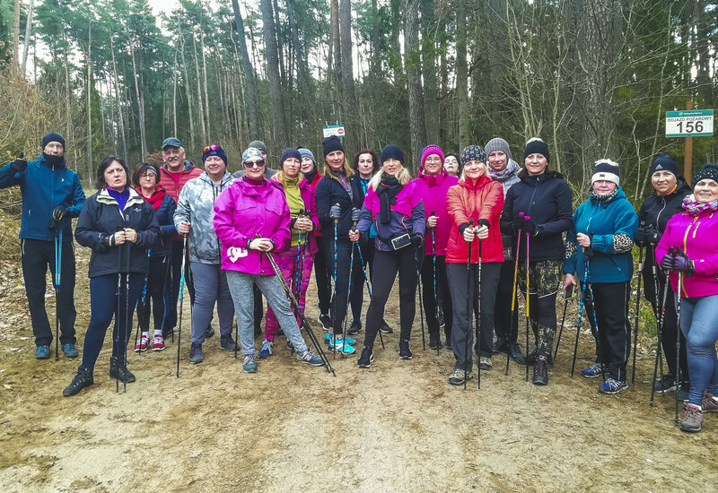 Grupa spacerujących w jasnych kurtkach i zimowych czapkach trzyma kijki do nordic walking na leśnej ścieżce, zrelaksowani i uśmiechnięci.