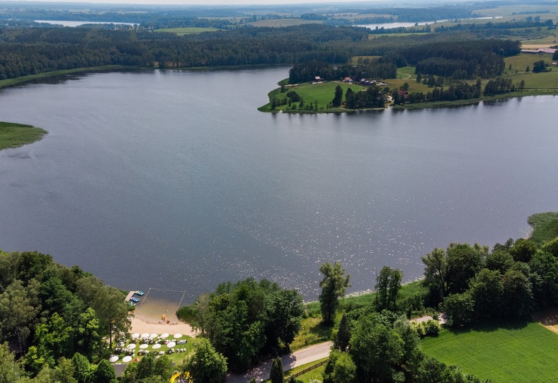 Widok z lotu ptaka na jezioro z hotelową piaszczystą plażą, parasolami, wypożyczalnią łodzi i zalesionym brzegiem