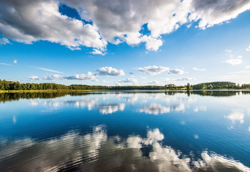 Spokojny widok na jezioro z lustrzaną taflą odbijającą chmury i zalesiony brzeg — idealny na relaks przy wodzie.