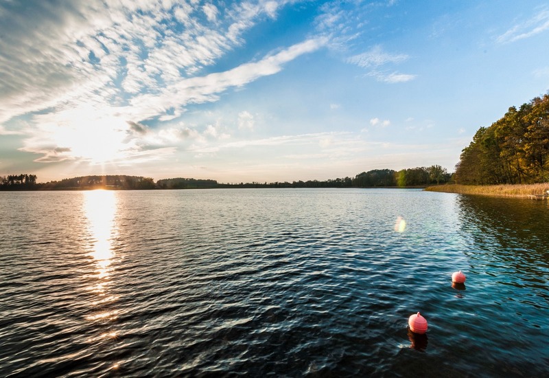 Zachód słońca nad spokojnym jeziorem, zalesiony brzeg, dwie pływające boje i połyskująca tafla — relaksujący widok.