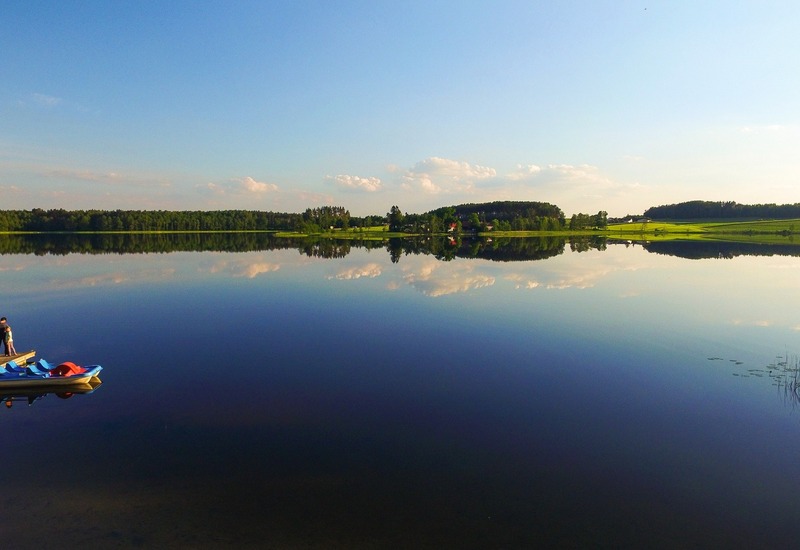 Spokojne jezioro z lustrzaną taflą, drewniany pomost z rowerkami wodnymi i grupą gości, zielone łąki i zalesione brzegi.