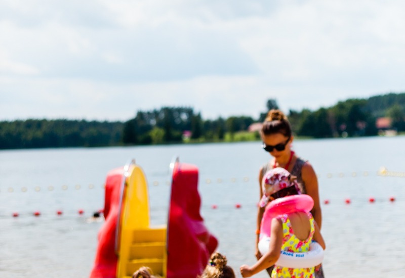 Plaża nad jeziorem z dziećmi w kolorowych kostiumach i dmuchanymi kołami, czerwono‑żółta zjeżdżalnia pedałowa, opiekun w pobliżu.