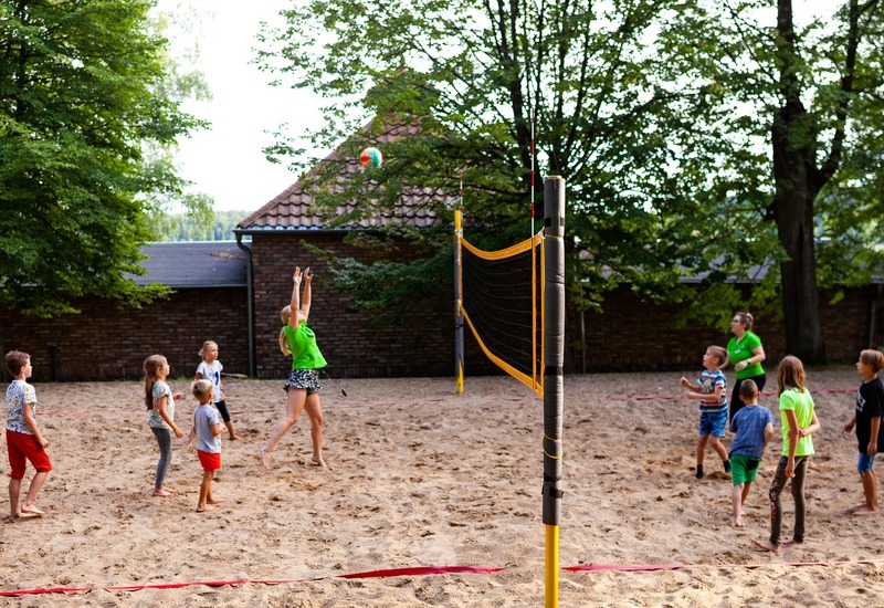 Dzieci z instruktorką grają w siatkówkę plażową na zacienionym boisku z piaskiem obok ceglanego budynku