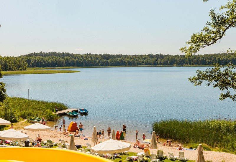 Piaszczysta plaża hotelowa nad spokojnym jeziorem z leżakami i parasolami, rodziny w wodzie, rowery wodne i pomost przy trzcinowym brzegu.