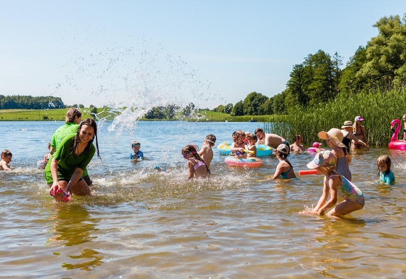 Rodzinna plaża nad jeziorem: dzieci i dorośli pluskają się w płytkiej wodzie, kolorowe dmuchane koła, trawiasty brzeg i drzewa.
