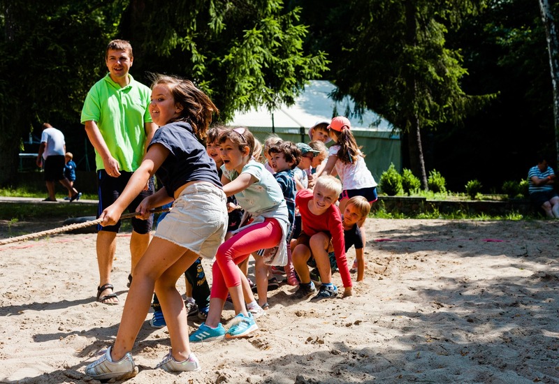Dzieci z animatorem przeciągają linę na piaszczystym boisku przy sosnach, nadzorowany klub dziecięcy