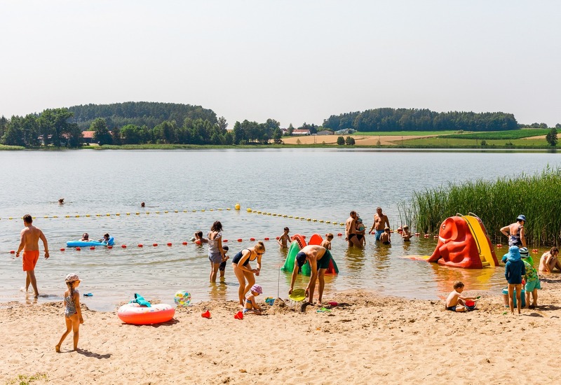 Rodzinna plaża nad jeziorem z wydzielonym kąpieliskiem, płytką wodą, dmuchaną zjeżdżalnią i bawiącymi się dziećmi.