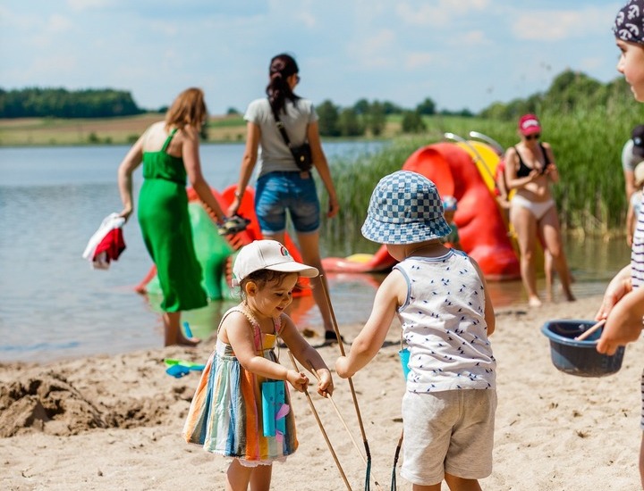 Piaszczysta plaża nad jeziorem w ośrodku rodzinnym; dzieci bawią się wiaderkami i siatkami, pontony i dorośli brodzą w wodzie