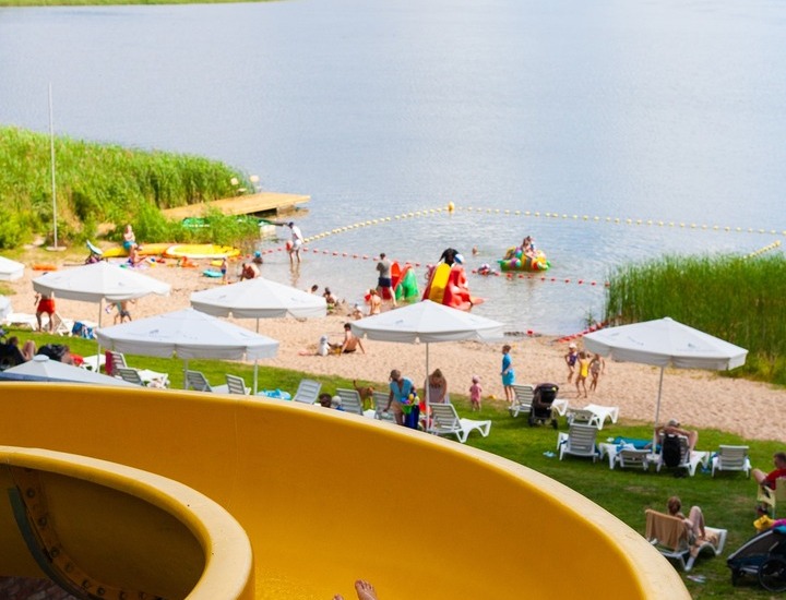 Dziecko na żółtej zjeżdżalni nad rodzinną plażą nad jeziorem z parasolami, leżakami i dmuchańcami.