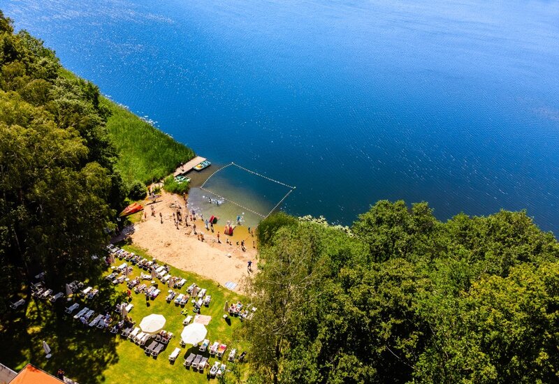 Piaszczysta plaża nad jeziorem z wyznaczonym kąpieliskiem, trawiasty taras z leżakami i parasolami, kajaki i bawiący się goście.