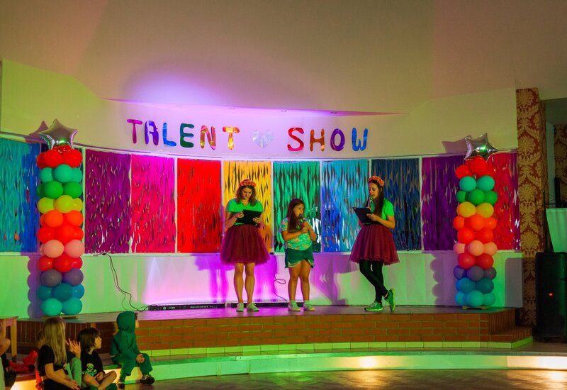Scena w sali konferencyjnej z banerem 'Talent Show', tęczowymi kolumnami z balonów, kolorowym tłem i dziećmi występującymi przy dekoracyjnym oświetleniu.