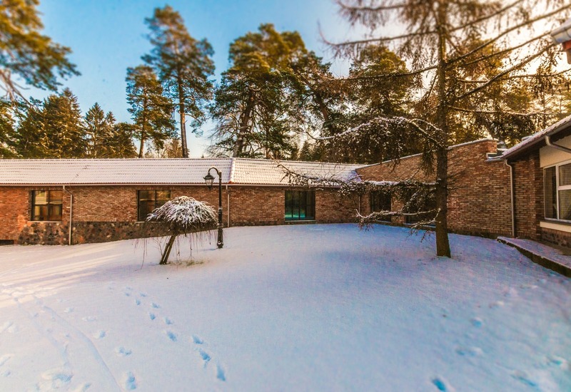 Zaśnieżony dziedziniec hotelu z niskim ceglanym budynkiem, latarnią, śladami stóp na śniegu, małymi drzewkami i wysokimi sosnami w tle.