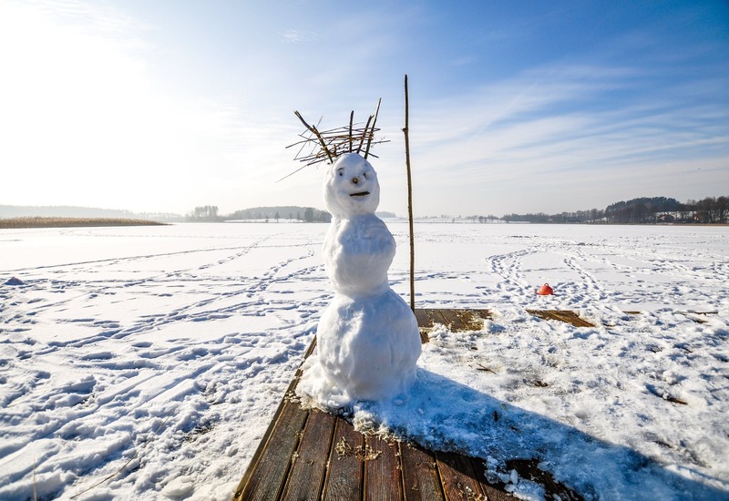 Bałwan z koroną z gałązek na drewnianym pomoście nad zamarzniętym jeziorem, błękitne niebo i ślady w śniegu
